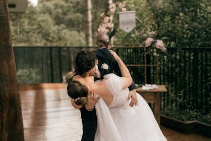 Newlywed couple sharing a romantic kiss outdoors on a wooden deck with lush greenery.