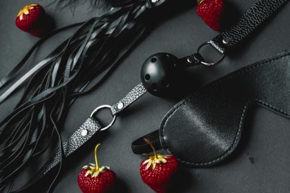 Flat lay of black leather BDSM items with fresh strawberries on dark background.