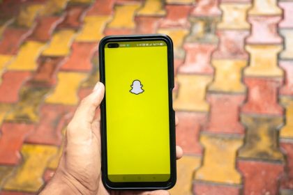 A person holds a smartphone displaying the Snapchat logo against a colorful brick background.