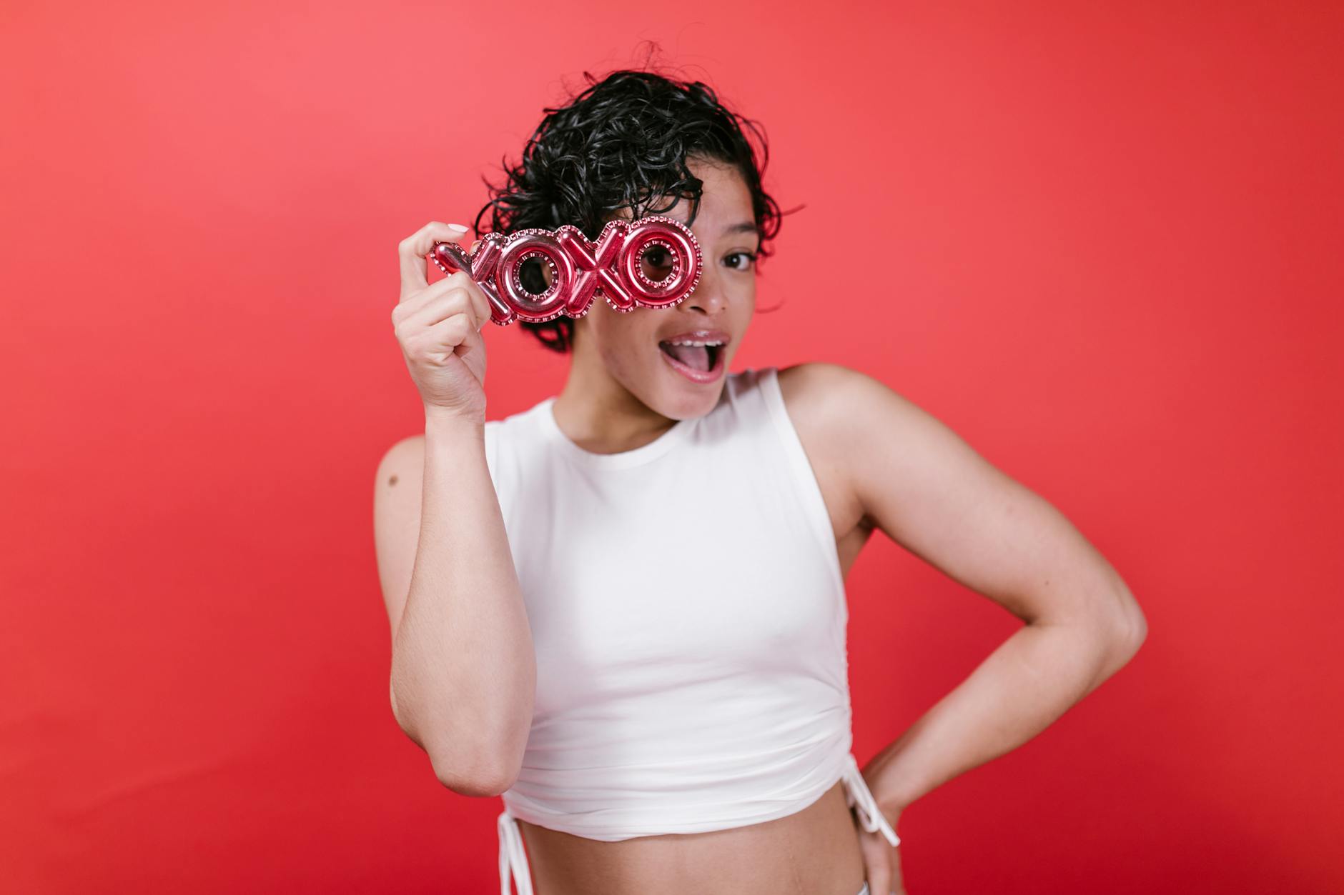 Happy woman in a white tank top holding XOXO glasses against a red background, symbolizing Valentine's Day joy.