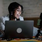Happy woman vlogging with a laptop and smartphone setup, creating content indoors.