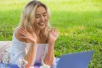 Young woman in grass making a video call, smiling and waving at the laptop camera.