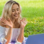 Young woman in grass making a video call, smiling and waving at the laptop camera.
