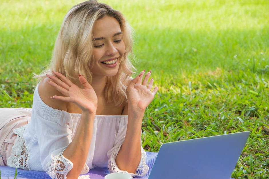 Young woman in grass making a video call, smiling and waving at the laptop camera.