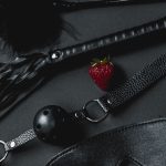 Un ensemble d'accessoires BDSM en cuir noir comprenant une balle de gag et un fouet, contrasté par une fraise fraîche.