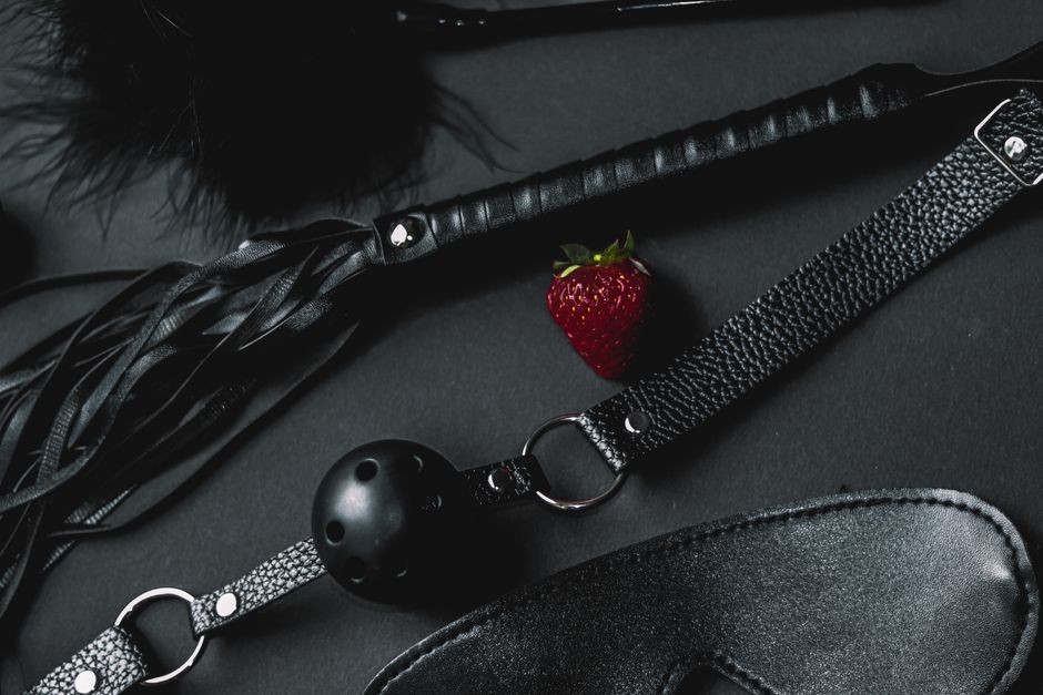 Un ensemble d'accessoires BDSM en cuir noir comprenant une balle de gag et un fouet, contrasté par une fraise fraîche.