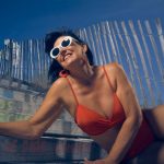 Une femme en bikini rouge sourit au soleil, appuyée contre une barrière en bois sur la plage.